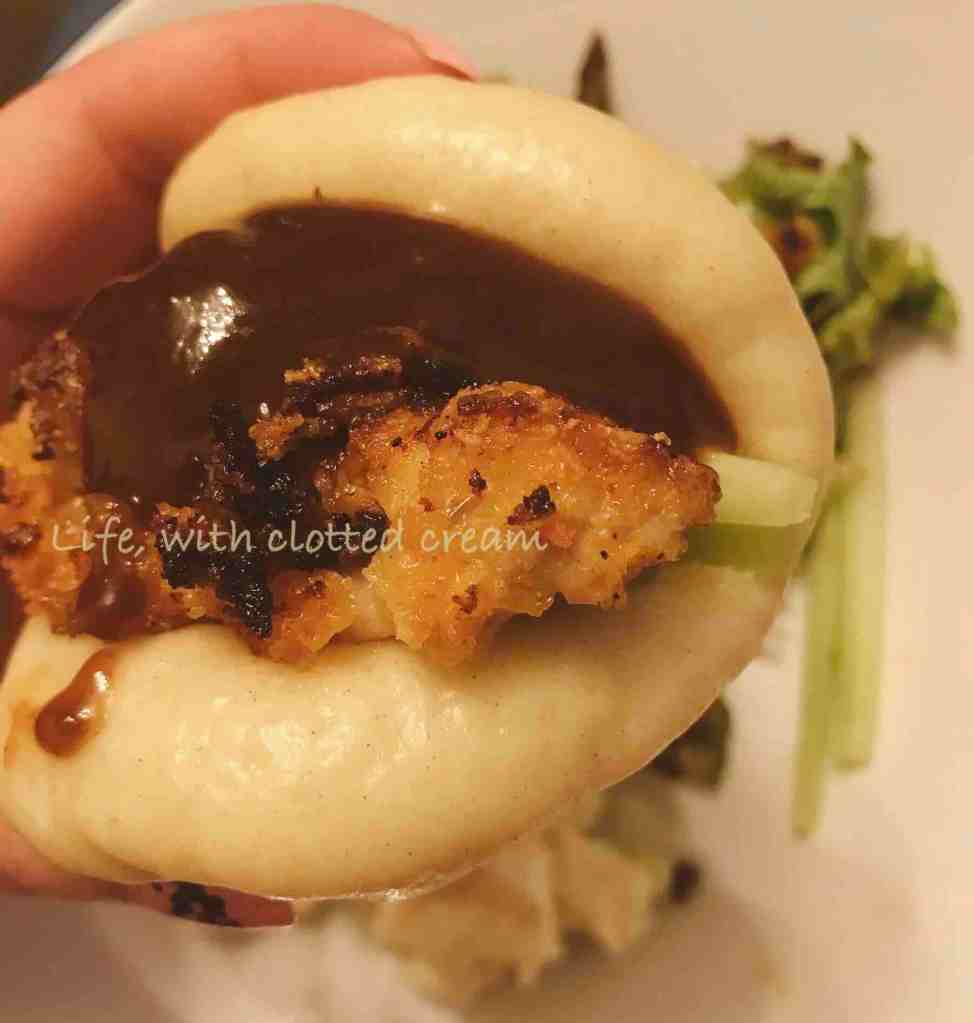 A bao with crispy chicken and hoisin sauce dripping towards the bottom. There is salad in the background.