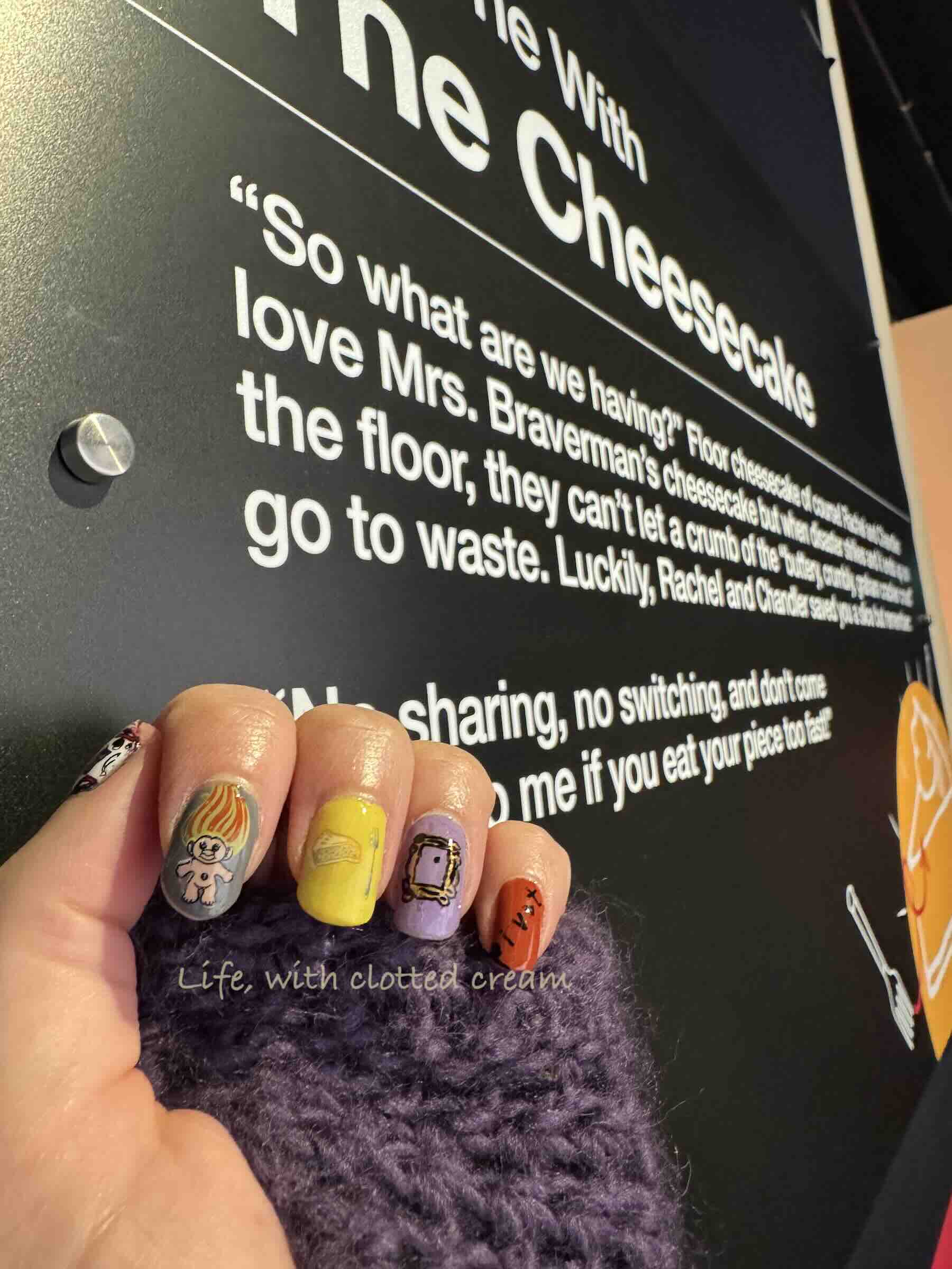 A board explaining the scene of the One with the Cheesecake. There is a hand in front with nails painted with Friends.