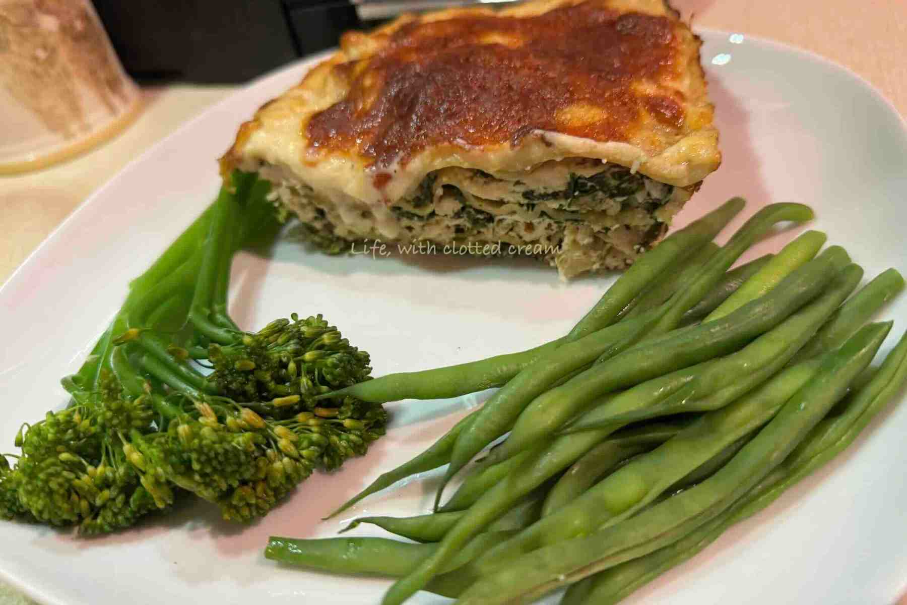 White chicken lasagna with golden crust and tenderstem broccoli and green beans on a white plate.