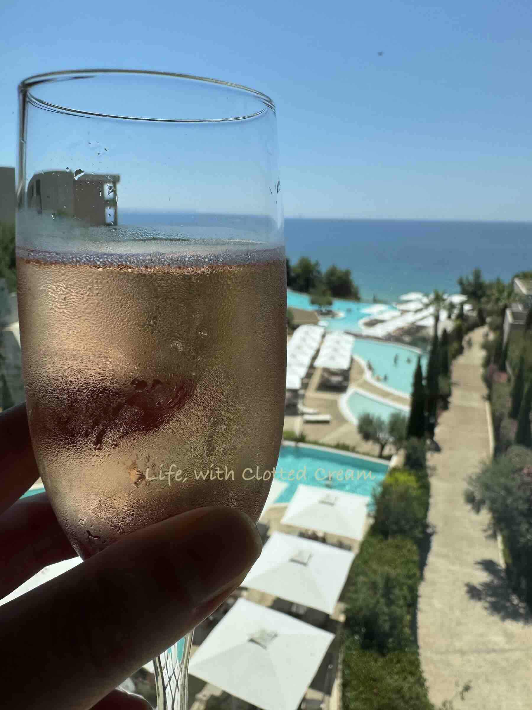A champagne glass with pink champagne overlooking swimming pools and the ocean.
