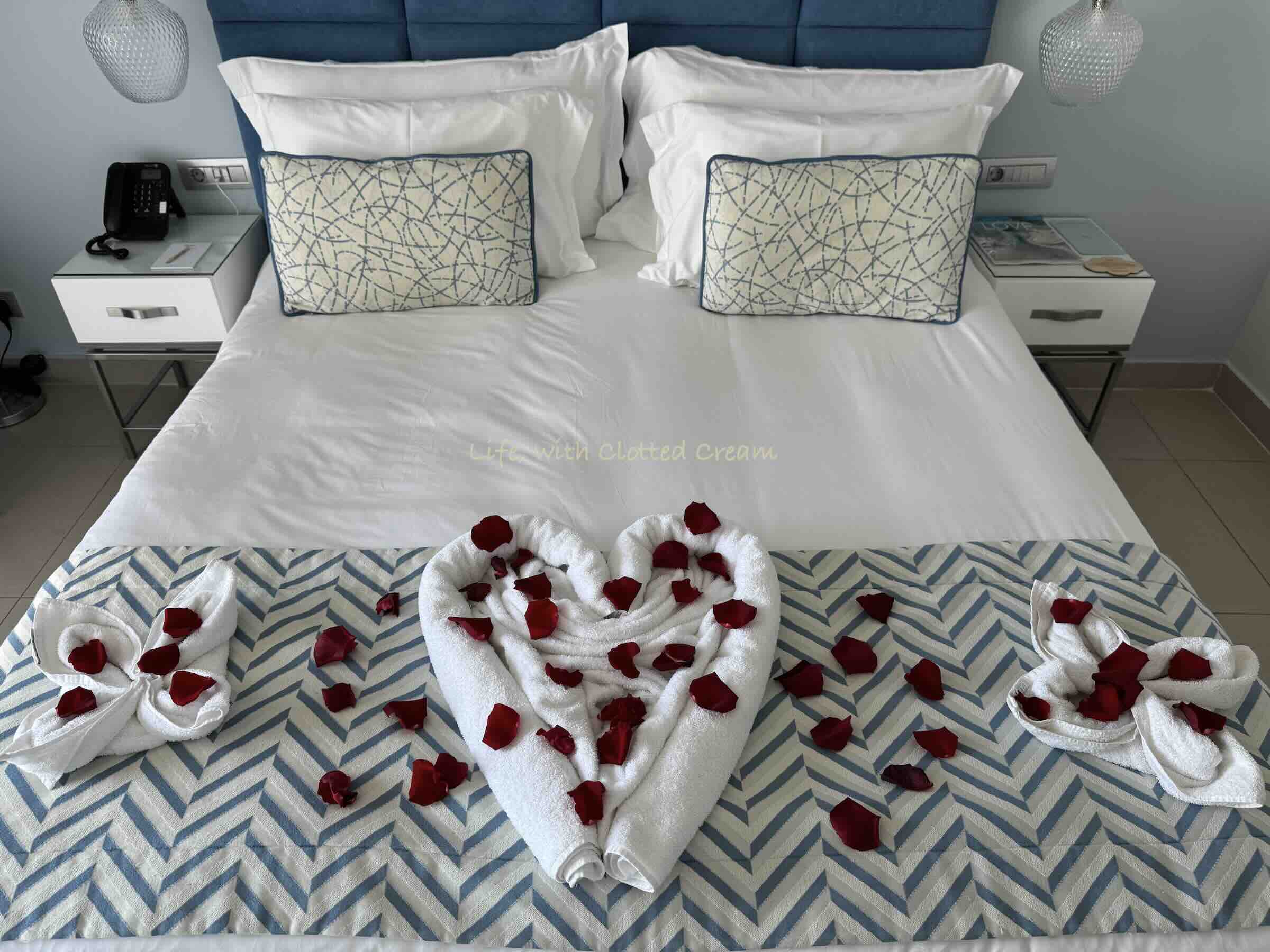 Double bed with towels arranged like hearts and red rose petals scattered around.