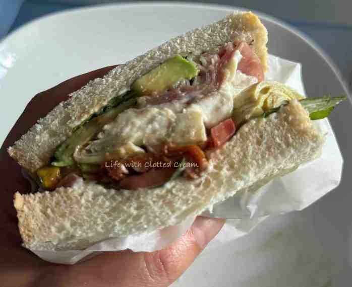 Sandwich wrapped in greaseproof paper held in a hand over a plate.